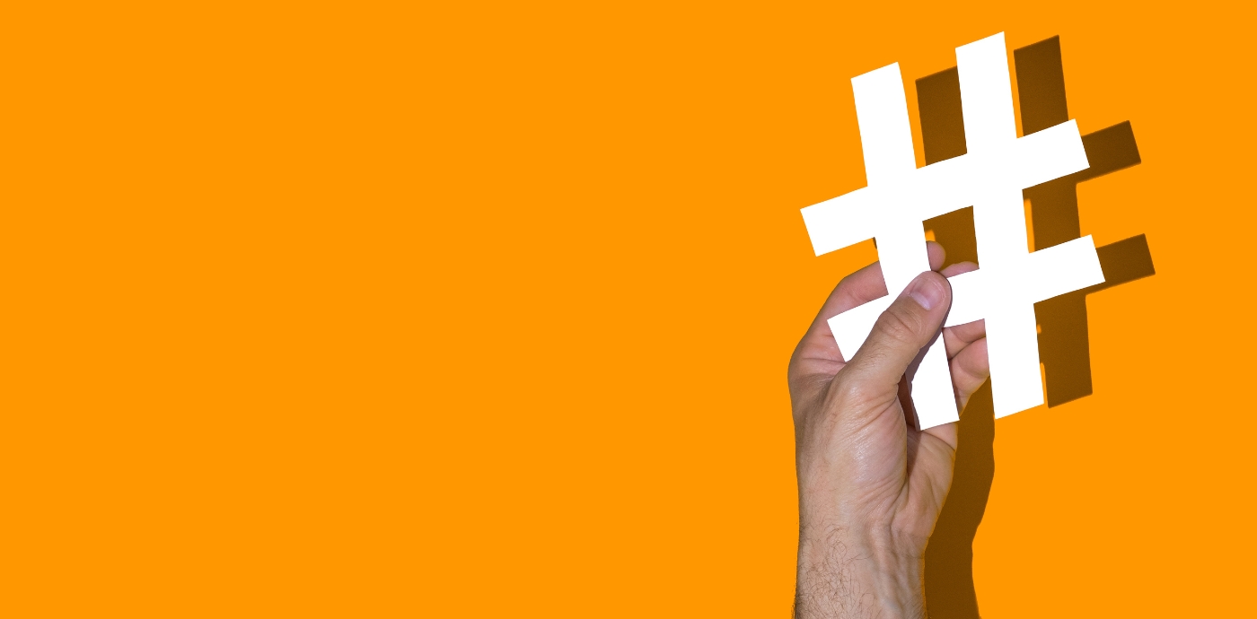 Person's hand holding up a paper cut out of a hash symbol against an orange background